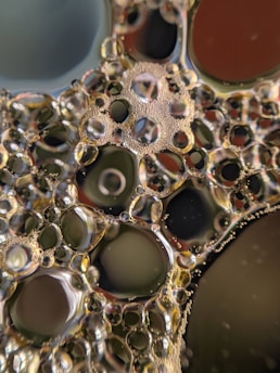 A detailed view of numerous oil droplets suspended in water, creating a mosaic of circular and oval shapes. The droplets range in size, with some containing smaller bubbles inside. Light reflects off the surface, creating a shimmering effect. The backdrop is a blend of muted colors, adding depth and contrast to the scene.