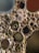 A detailed view of numerous oil droplets suspended in water, creating a mosaic of circular and oval shapes. The droplets range in size, with some containing smaller bubbles inside. Light reflects off the surface, creating a shimmering effect. The backdrop is a blend of muted colors, adding depth and contrast to the scene.