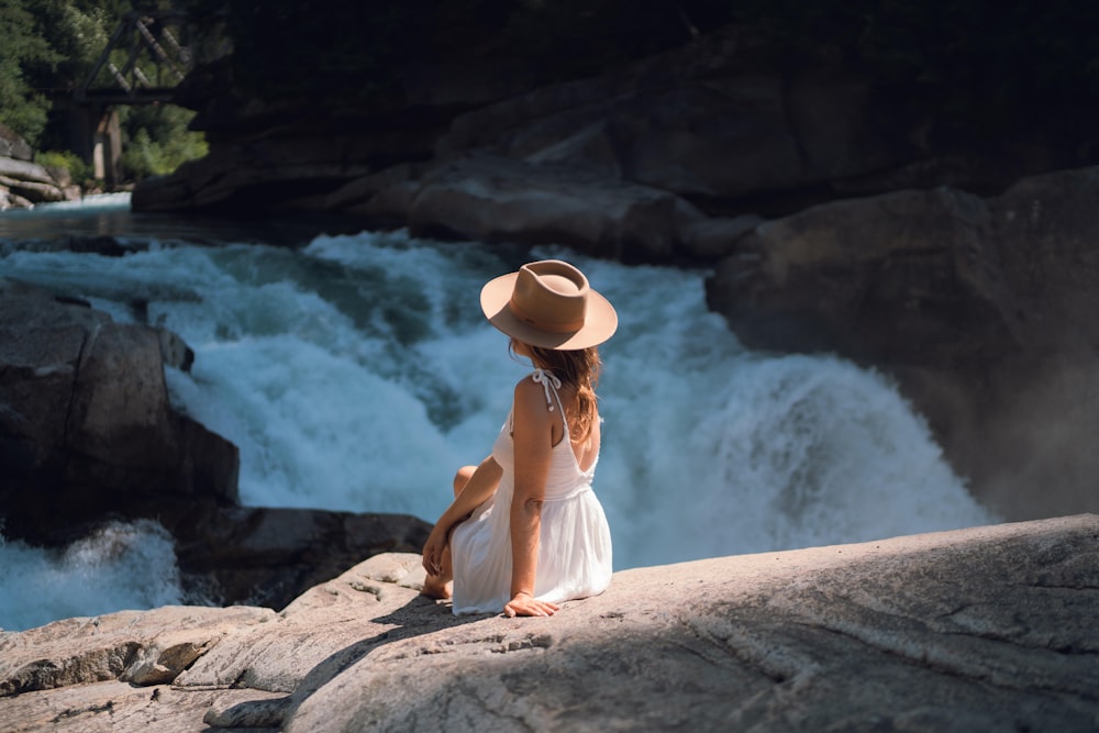 woman in white dress sitting on rock near water falls during daytime