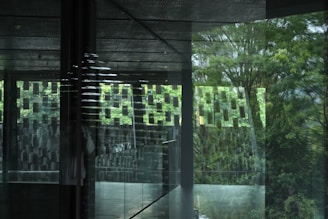 A modern architectural space featuring large glass panels reflecting an outdoor scene with lush green trees. The interior has geometric patterns on the walls, creating an abstract and artistic atmosphere.