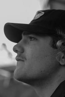 A black and white profile portrait of a person wearing a hat. The focus is on the face, showing a thoughtful expression with subtle details like facial hair and curly hair peeking out from under the cap.