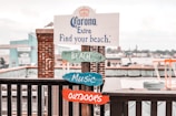 A wooden signpost stands by a waterfront, featuring a promotional message for Corona Extra with the phrase 'Find your beach.' Underneath, colorful wooden signs labeled 'BEACH,' 'MUSIC,' and 'OUTDOORS' are attached. The backdrop includes a brick pillar and an industrial or urban waterfront area with buildings and boats.