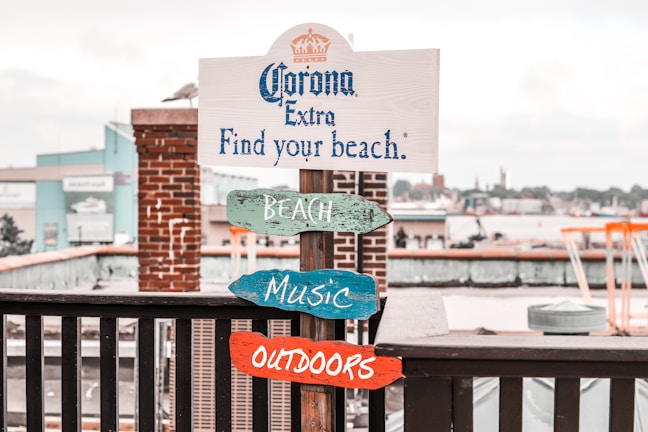 A wooden signpost stands by a waterfront, featuring a promotional message for Corona Extra with the phrase 'Find your beach.' Underneath, colorful wooden signs labeled 'BEACH,' 'MUSIC,' and 'OUTDOORS' are attached. The backdrop includes a brick pillar and an industrial or urban waterfront area with buildings and boats.