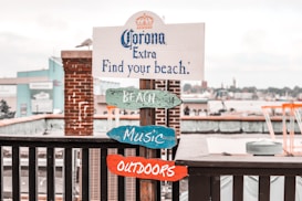 A wooden signpost stands by a waterfront, featuring a promotional message for Corona Extra with the phrase 'Find your beach.' Underneath, colorful wooden signs labeled 'BEACH,' 'MUSIC,' and 'OUTDOORS' are attached. The backdrop includes a brick pillar and an industrial or urban waterfront area with buildings and boats.