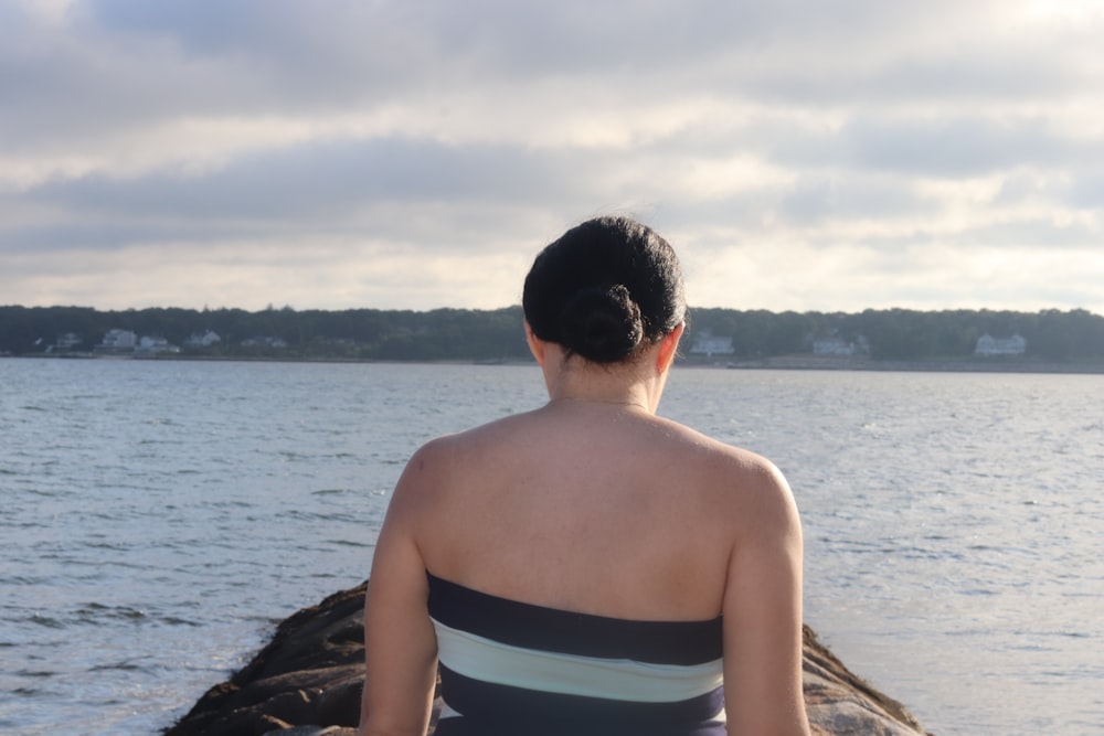 woman in white and blue striped tube dress sitting on brown rock near body of water