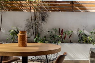 A minimalist outdoor setting features a round wooden table with a decorative ceramic pot in the center. The background includes a planter with various green and red-leafed plants against a white wall, partially shaded by a wooden slat fence.