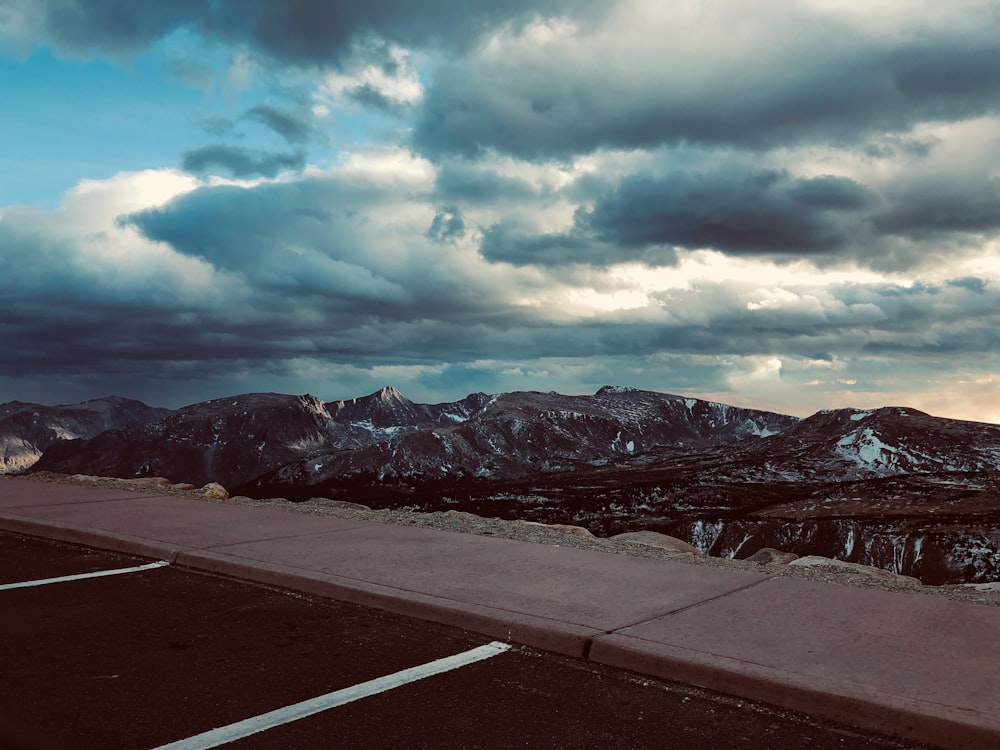 gray concrete road near snow covered mountains under cloudy sky during daytime