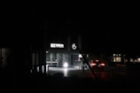A dimly lit entrance to a building with a glowing 'UIC Service City Centre' sign and a circular parking symbol above the doorway. There is a car with its rear lights on approaching the entrance. The surroundings are dark, with minimal lighting in the background and some foliage visible.
