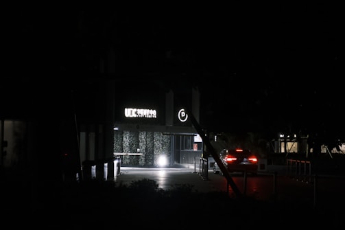 A dimly lit entrance to a building with a glowing 'UIC Service City Centre' sign and a circular parking symbol above the doorway. There is a car with its rear lights on approaching the entrance. The surroundings are dark, with minimal lighting in the background and some foliage visible.