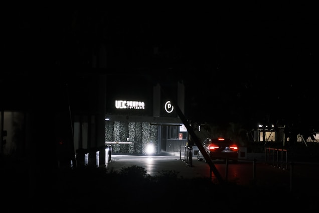 A dimly lit entrance to a building with a glowing 'UIC Service City Centre' sign and a circular parking symbol above the doorway. There is a car with its rear lights on approaching the entrance. The surroundings are dark, with minimal lighting in the background and some foliage visible.
