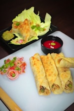 A plate of neatly arranged stuffed wraps with a golden-brown finish. The wraps are accompanied by a side of fresh vegetable salad consisting of sliced onions, tomatoes, and herbs. In the background, a dish of crispy fried items rests on a bed of lettuce with a green dip. A small bowl filled with bright red sauce is also visible on the side.