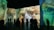 A large room featuring immersive digital projections of impressionist-style artwork focusing on close-up details of faces. Silhouettes of people are seen observing the vibrant and expressive paintings, with various postures suggesting engagement with the art.