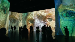 A large room featuring immersive digital projections of impressionist-style artwork focusing on close-up details of faces. Silhouettes of people are seen observing the vibrant and expressive paintings, with various postures suggesting engagement with the art.