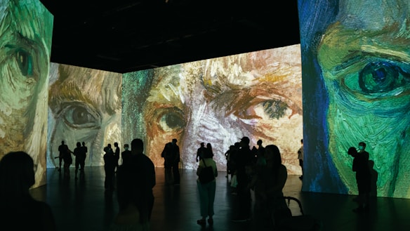 A large room featuring immersive digital projections of impressionist-style artwork focusing on close-up details of faces. Silhouettes of people are seen observing the vibrant and expressive paintings, with various postures suggesting engagement with the art.