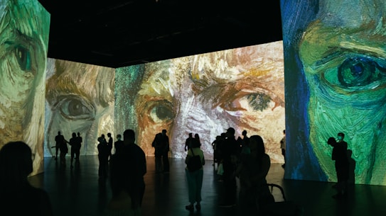 A large room featuring immersive digital projections of impressionist-style artwork focusing on close-up details of faces. Silhouettes of people are seen observing the vibrant and expressive paintings, with various postures suggesting engagement with the art.