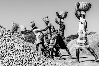 A group of men wearing traditional attire are working together to move a large pile of stones. Each man carries a basket, either lifting it high above their heads or pouring its contents onto the pile. The scene is captured in black and white, highlighting their muscular effort and teamwork under the clear sky.