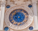 A detailed and ornate astronomical clock with blue and gold tones, featuring zodiac symbols and Roman numerals. The clock is set within a stone facade with two small circular windows on either side.