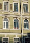 The image depicts a section of an ornate, historic building facade with several windows. The building is painted in a muted green color with white decorative trim. There are intricate architectural details above and between the windows. Inside one of the windows, a person wearing a bright orange outfit and a helmet is visible, possibly engaged in work.