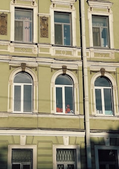 The image depicts a section of an ornate, historic building facade with several windows. The building is painted in a muted green color with white decorative trim. There are intricate architectural details above and between the windows. Inside one of the windows, a person wearing a bright orange outfit and a helmet is visible, possibly engaged in work.
