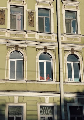 The image depicts a section of an ornate, historic building facade with several windows. The building is painted in a muted green color with white decorative trim. There are intricate architectural details above and between the windows. Inside one of the windows, a person wearing a bright orange outfit and a helmet is visible, possibly engaged in work.