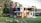 A modern two-story house with a sleek design featuring a mix of wood and glass elements. The facade includes large windows and balconies with glass railings. The surroundings are lush with well-maintained grass, rocks, and various trees.