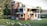 A modern two-story house with a sleek design featuring a mix of wood and glass elements. The facade includes large windows and balconies with glass railings. The surroundings are lush with well-maintained grass, rocks, and various trees.