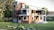 A modern two-story house with a sleek design featuring a mix of wood and glass elements. The facade includes large windows and balconies with glass railings. The surroundings are lush with well-maintained grass, rocks, and various trees.