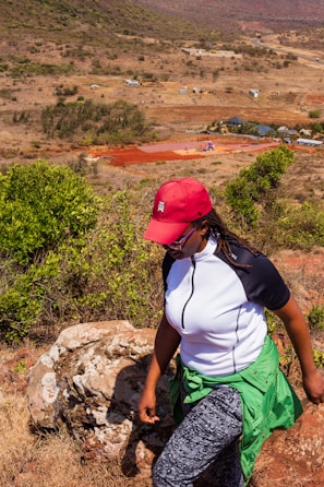 A person wearing a red cap and white and black shirt is hiking on a rocky trail. The landscape is dry with sparse vegetation, featuring a vast, open area with a few buildings and a winding road in the background.