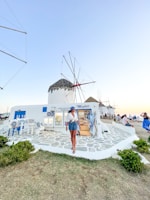 A person is walking along a path in front of a white building displaying Greek traditional products. The building has blue accents, matching the outdoor decor, and features a thatched-roof windmill in the background. Other people are sitting nearby, enjoying the scenery. The sky is clear, suggesting a peaceful atmosphere.