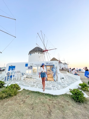 A person is walking along a path in front of a white building displaying Greek traditional products. The building has blue accents, matching the outdoor decor, and features a thatched-roof windmill in the background. Other people are sitting nearby, enjoying the scenery. The sky is clear, suggesting a peaceful atmosphere.