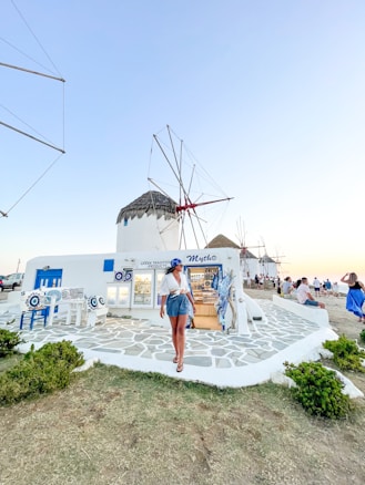 A person is walking along a path in front of a white building displaying Greek traditional products. The building has blue accents, matching the outdoor decor, and features a thatched-roof windmill in the background. Other people are sitting nearby, enjoying the scenery. The sky is clear, suggesting a peaceful atmosphere.