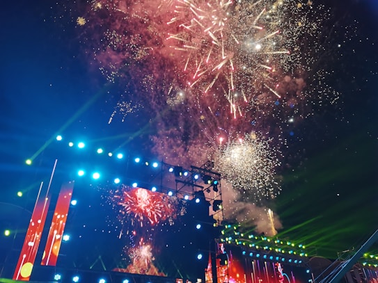 A vibrant display of fireworks lights up the night sky above a large stage adorned with bright lights and screens. The fireworks burst in various colors, including red, white, and yellow, creating a festive atmosphere. The stage itself is illuminated with multicolored lights, adding to the celebratory feel of the scene.