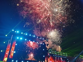 A vibrant display of fireworks lights up the night sky above a large stage adorned with bright lights and screens. The fireworks burst in various colors, including red, white, and yellow, creating a festive atmosphere. The stage itself is illuminated with multicolored lights, adding to the celebratory feel of the scene.