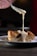 A rich and creamy sauce is slowly being poured from a spoon onto a piece of bread pudding. The dessert sits on a white plate, with a backdrop that is slightly out of focus, highlighting the decadent texture and inviting appearance of the dish.