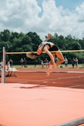 A high jumper gracefully clears the bar during a track and field event. The athlete is in mid-air, arching their back over the bar with precision. The background features lush green trees and a partly cloudy sky, with spectators visible in the distance. The track surface and landing mat are prominently displayed, with vivid orange-red and pink hues.