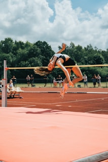 A high jumper gracefully clears the bar during a track and field event. The athlete is in mid-air, arching their back over the bar with precision. The background features lush green trees and a partly cloudy sky, with spectators visible in the distance. The track surface and landing mat are prominently displayed, with vivid orange-red and pink hues.