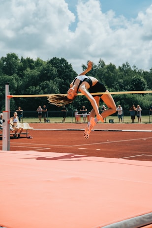 A high jumper gracefully clears the bar during a track and field event. The athlete is in mid-air, arching their back over the bar with precision. The background features lush green trees and a partly cloudy sky, with spectators visible in the distance. The track surface and landing mat are prominently displayed, with vivid orange-red and pink hues.