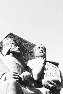 Two statues of bearded men are depicted, carved from stone in a classical style. One figure appears to be holding a scroll or tablet featuring inscriptions, while the other holds an orb. The statues exude a sense of wisdom and history, positioned against a clear sky.