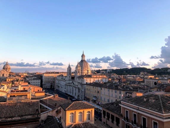 A panoramic view of a cityscape during golden hour, showcasing classic European architecture with prominent domes and terracotta rooftops. The sky is clear with scattered clouds, and the warm sunlight casts a soft glow on the buildings.
