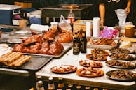 An array of grilled and roasted meats, including whole poultry, sausages, and various cuts, is elegantly displayed on trays and plates. Bottles of Guinness beer are also visible on the table. In the background, a grill can be seen along with utensils and containers, suggesting a vibrant food market or stall setting.