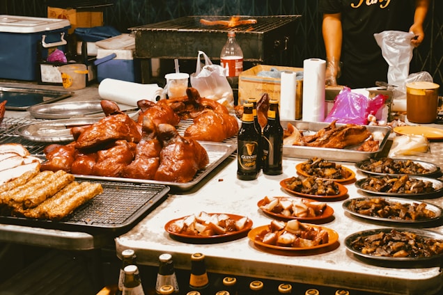An array of grilled and roasted meats, including whole poultry, sausages, and various cuts, is elegantly displayed on trays and plates. Bottles of Guinness beer are also visible on the table. In the background, a grill can be seen along with utensils and containers, suggesting a vibrant food market or stall setting.