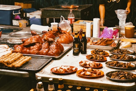 An array of grilled and roasted meats, including whole poultry, sausages, and various cuts, is elegantly displayed on trays and plates. Bottles of Guinness beer are also visible on the table. In the background, a grill can be seen along with utensils and containers, suggesting a vibrant food market or stall setting.