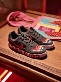 A pair of black sneakers with a red logo, featuring colorful designs on the toe area. The midsole has large white graphics, and the shoes are placed on a wooden surface. In the background, a pink cloth and some printed materials like magazines or booklets are visible.