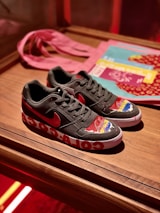 A pair of black sneakers with a red logo, featuring colorful designs on the toe area. The midsole has large white graphics, and the shoes are placed on a wooden surface. In the background, a pink cloth and some printed materials like magazines or booklets are visible.