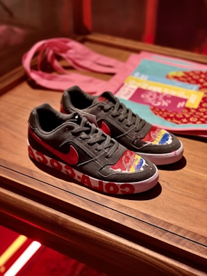 A pair of black sneakers with a red logo, featuring colorful designs on the toe area. The midsole has large white graphics, and the shoes are placed on a wooden surface. In the background, a pink cloth and some printed materials like magazines or booklets are visible.