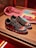 A pair of black sneakers with a red logo, featuring colorful designs on the toe area. The midsole has large white graphics, and the shoes are placed on a wooden surface. In the background, a pink cloth and some printed materials like magazines or booklets are visible.