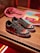 A pair of black sneakers with a red logo, featuring colorful designs on the toe area. The midsole has large white graphics, and the shoes are placed on a wooden surface. In the background, a pink cloth and some printed materials like magazines or booklets are visible.