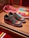 A pair of black sneakers with a red logo, featuring colorful designs on the toe area. The midsole has large white graphics, and the shoes are placed on a wooden surface. In the background, a pink cloth and some printed materials like magazines or booklets are visible.