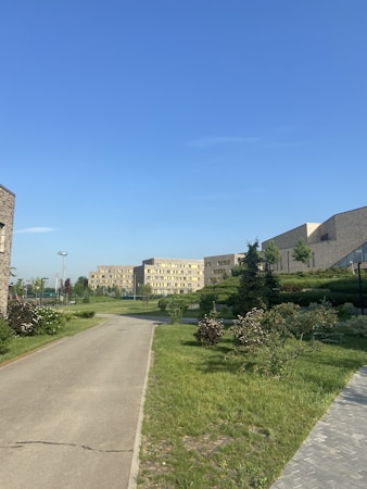 A pathway runs through a well-maintained green area with bushes and grass, flanked by modern buildings in the background. The sky is clear and bright blue, suggesting a calm weather.