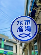 A round sign with blue Japanese characters and a stylized fish symbol against a white background is displayed outside. It is mounted on the side of a building, with an urban street scene partially visible in the background and a clear blue sky above.